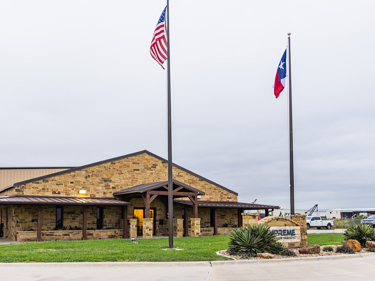 The Supreme Production Services headquarters on State Highway 44 in Corpus Christi, with the U.S. and Texas flags flying over the entrance and the company monument sign at the drive.